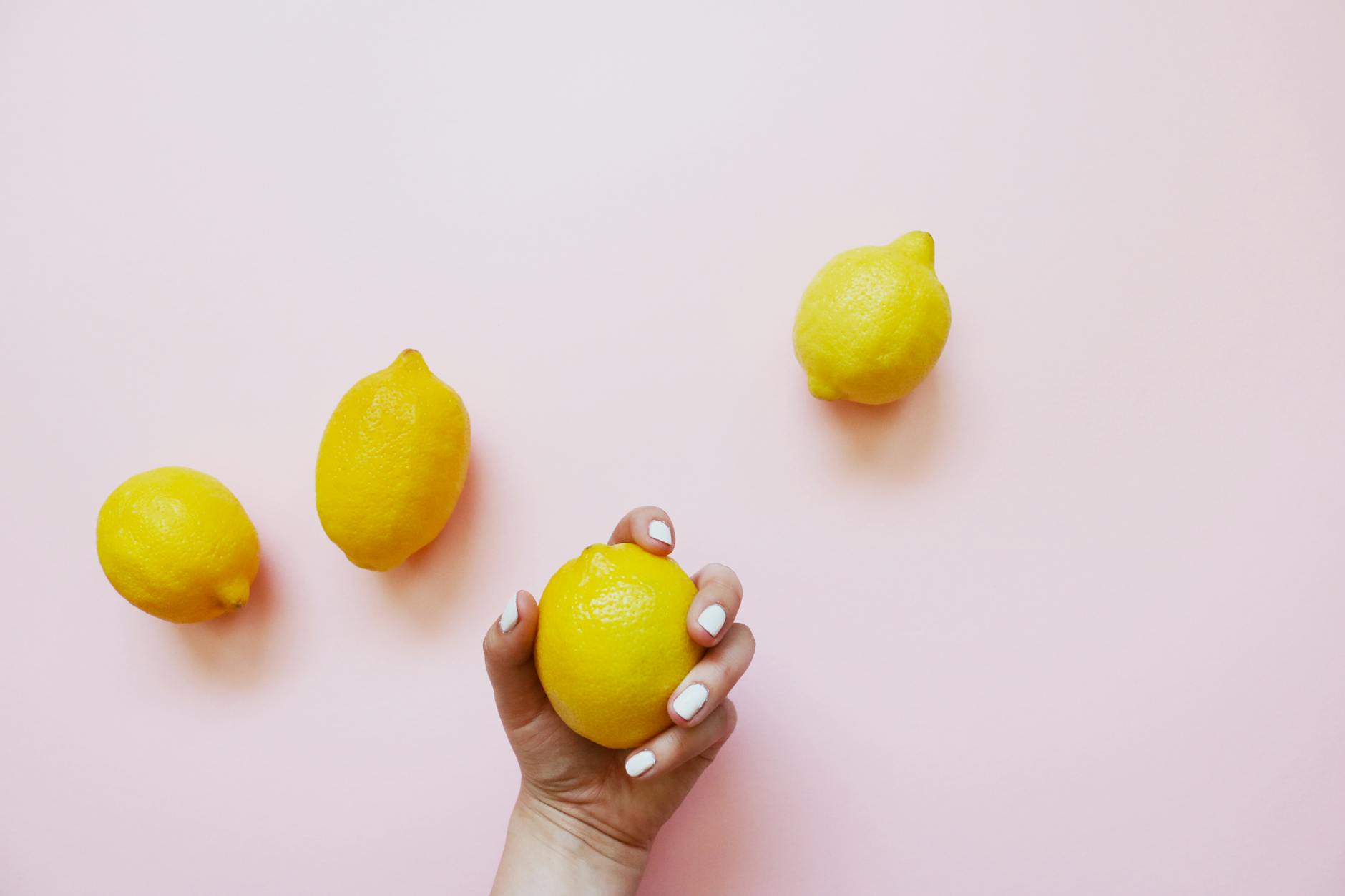 A hand with white nails holding a lemon on a soft pink background, surrounded by fresh lemons